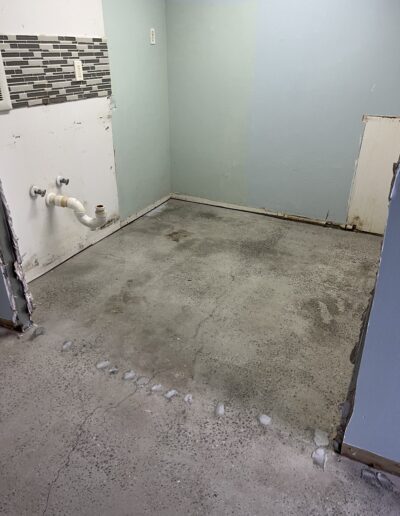 image shows a room with a bare concrete subfloor. There's a blue wall on the right and a light green wall at the back. A white door is leaning against the blue wall